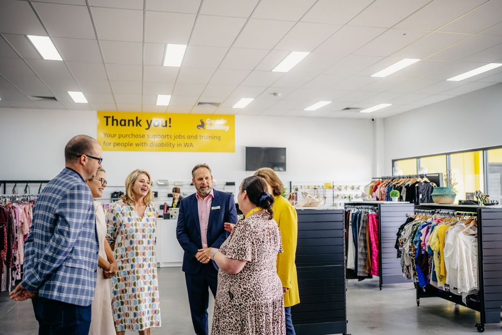 Group of people conversing inside a Good Sammy store beneath a yellow thank you sign.