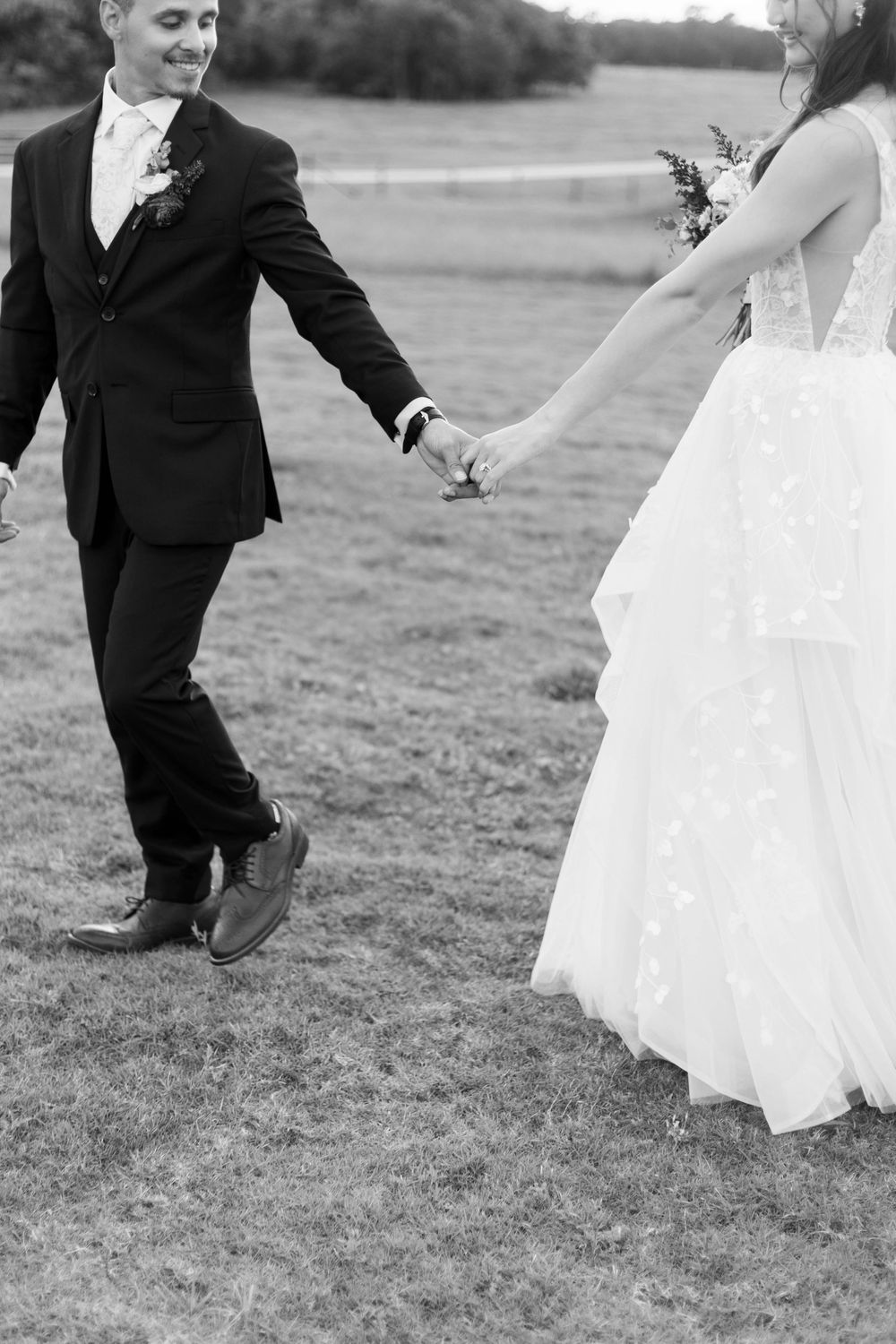 Black and white photo of bride and groom holding hands walking on grass, bride in lace gown, groom in dark suit.