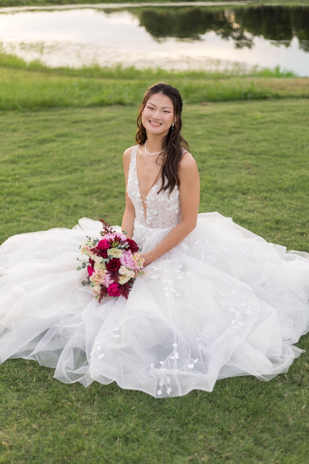 Smiling bride in white floral tulle gown sits on green grass holding pink bouquet near a serene lake.