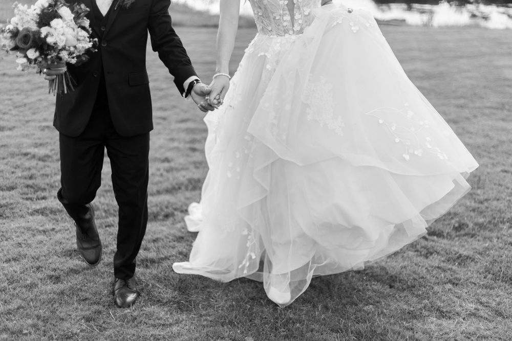 Black and white photo of bride and groom walking hand in hand on grass, bride holding bouquet in flowing wedding gown.