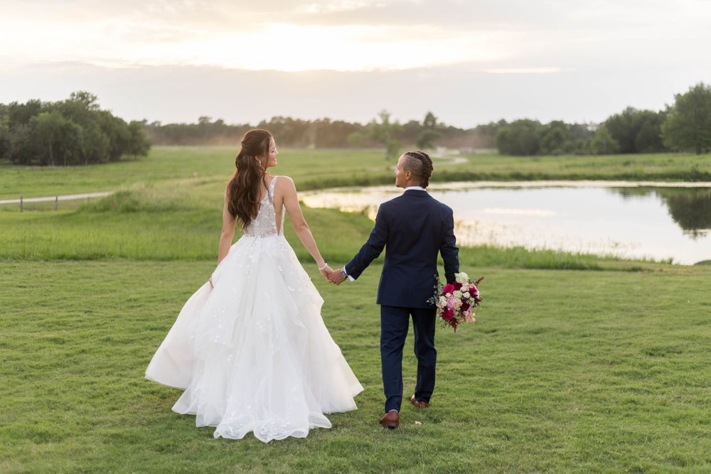 Bride and groom holding hands walking toward a scenic pond at sunset on their wedding day.
