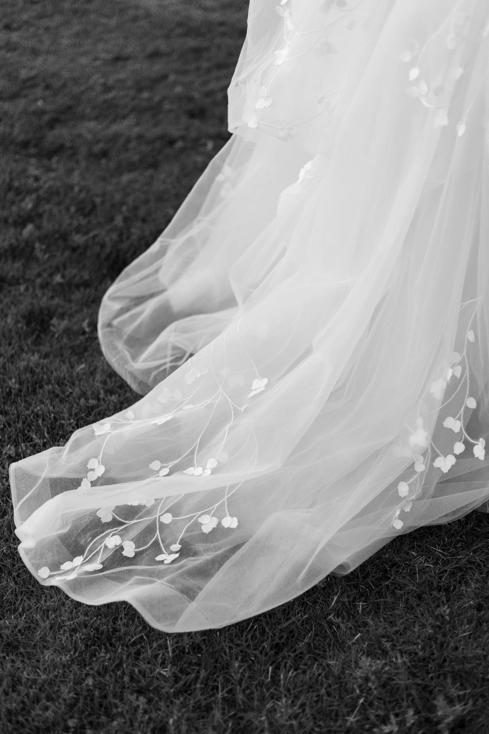 Black and white photo of a flowing bridal gown train with delicate floral embroidery on tulle, spread across green grass.