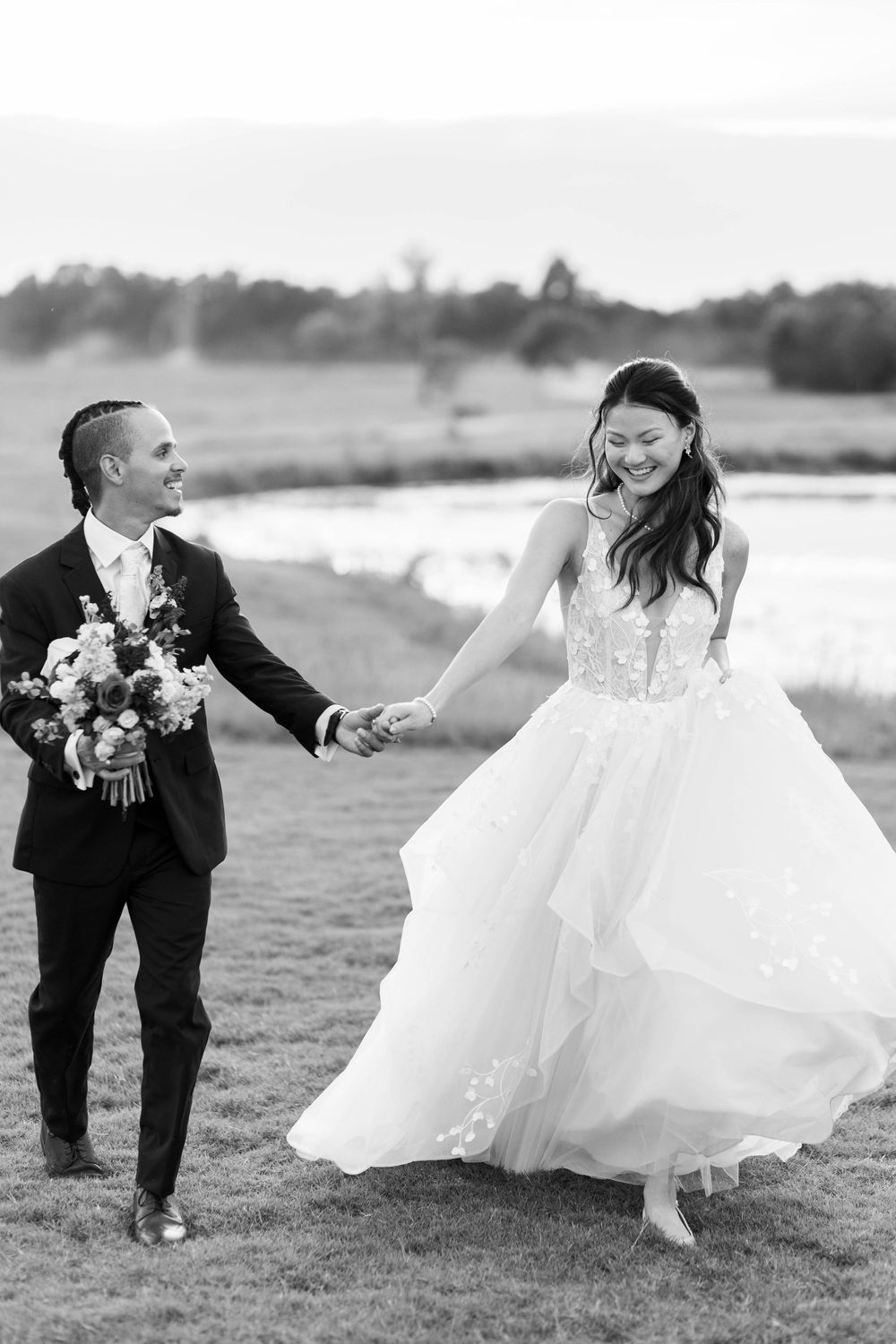Black and white photo of a smiling bride and groom holding hands while walking outdoors on their wedding day.