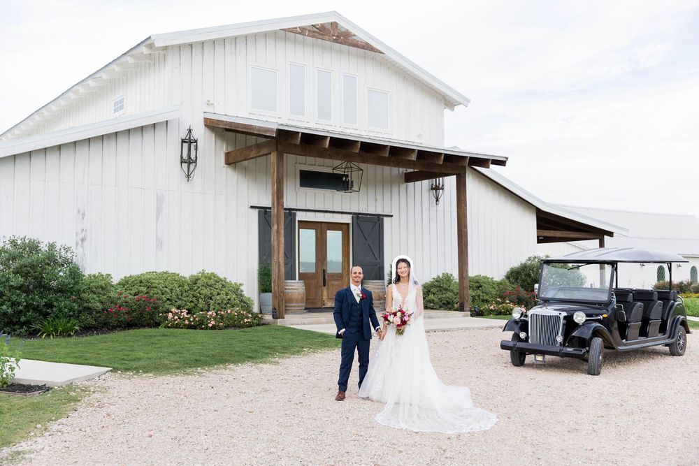 Bride and groom walk hand-in-hand outside a white barn wedding venue with a vintage golf cart nearby.