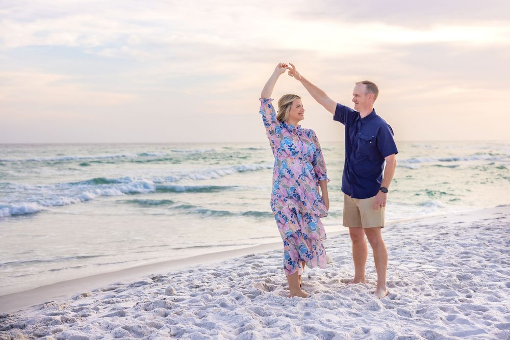 Older couple dancing joyfully on a sandy beach at sunset, waves and pastel sky in the background.