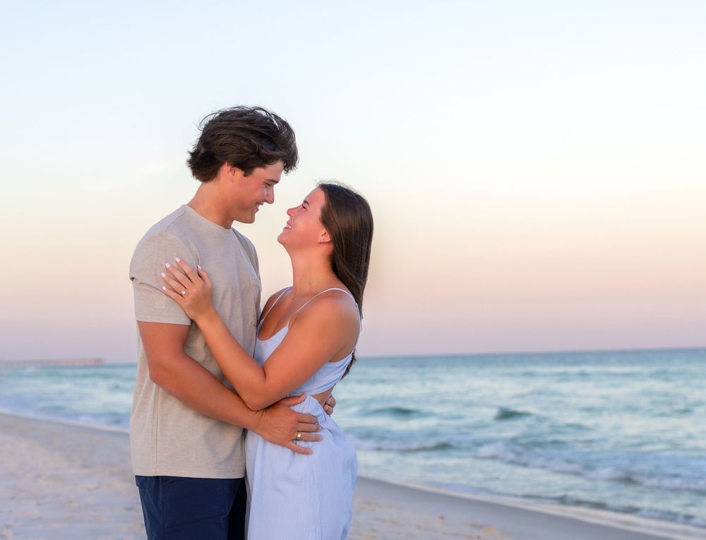 Romantic couple embracing on a sandy beach at sunset with calm ocean waves in the background.