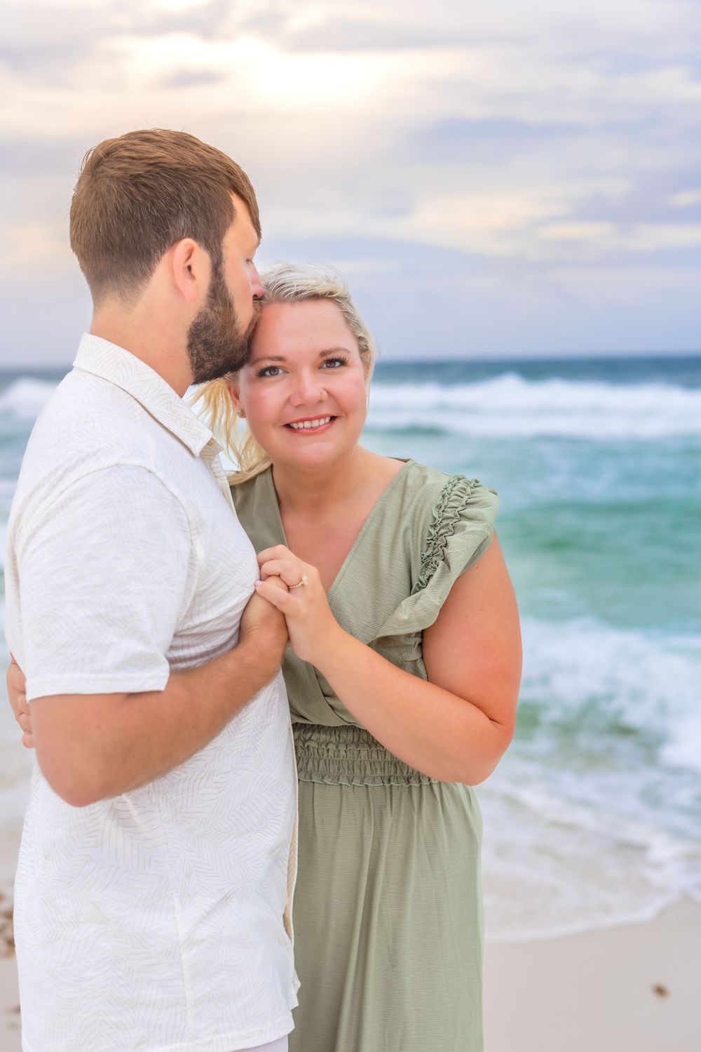 Couple embracing on a beach at sunset, woman in green dress smiling, turquoise ocean waves in the background.
