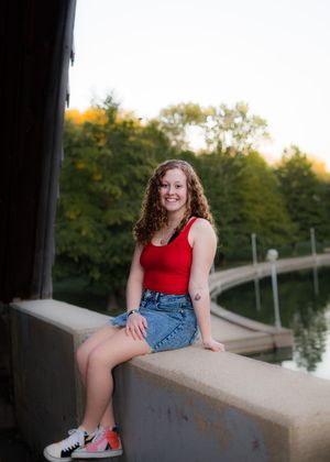 Young woman with curly hair sits on a ledge in a red tank top and denim skirt, smiling by a scenic waterway.