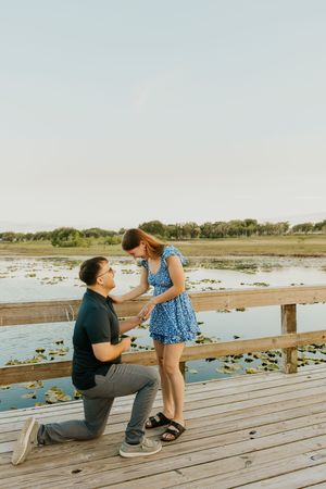 Man kneeling on wooden dock proposing to woman in blue dress by a scenic lake at sunset.