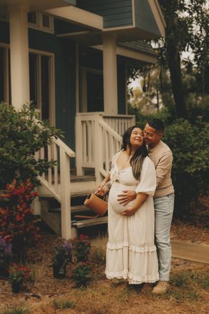 Couple embracing outdoors in front of blue house, pregnant woman in white dress held by partner in maternity photo shoot.