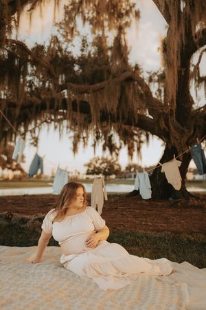 Pregnant woman in white dress sitting on blanket under Spanish moss oak tree with baby clothes hanging on a line.