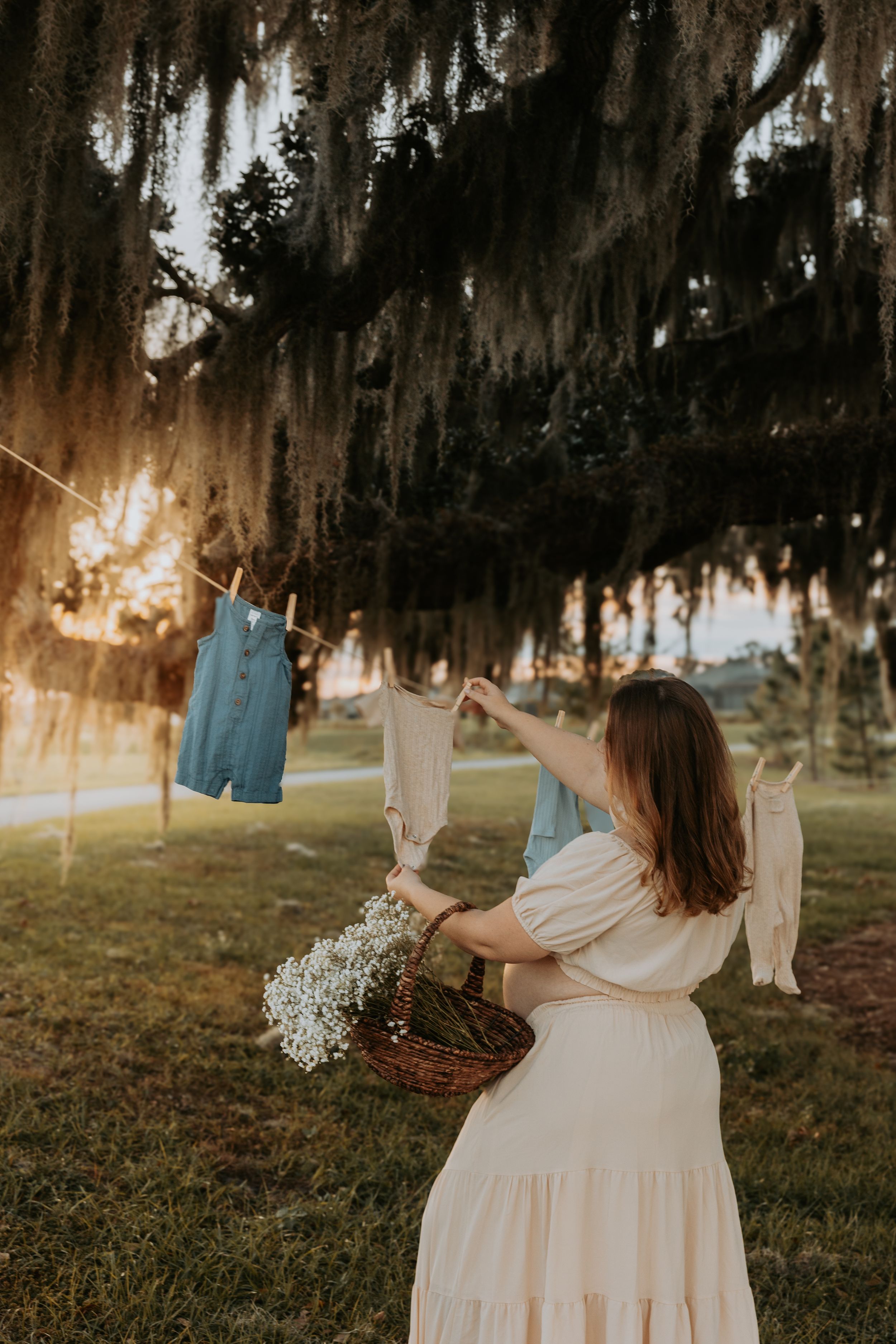 Woman in cream dress hanging baby clothes on a clothesline under moss-draped trees at golden hour.