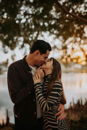 Couple sharing a romantic almost-kiss at golden hour by a lake, woman in striped sweater embraced by man in burgundy shirt.