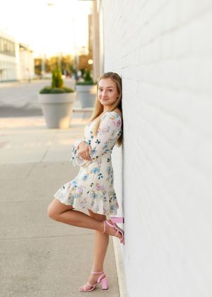 Young woman in floral mini dress and pink heels posing against white brick wall outdoors at golden hour.