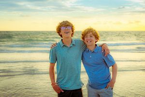 Two smiling teenage boys with curly hair pose together on a beach at sunset wearing blue polo shirts.