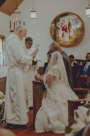Bride and groom kneel before a priest during a traditional church wedding ceremony with stained glass backdrop.