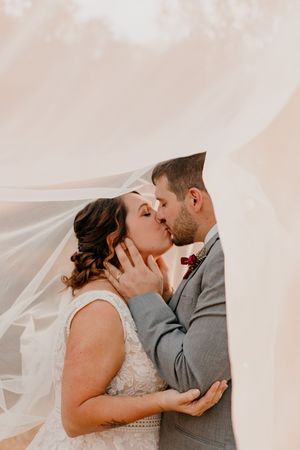 Bride and groom sharing a romantic kiss under a flowing wedding veil in a dreamy outdoor portrait.