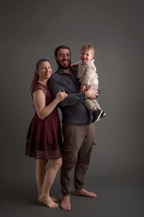 A smiling family of three poses in a studio, with parents holding their laughing toddler against a gray background.