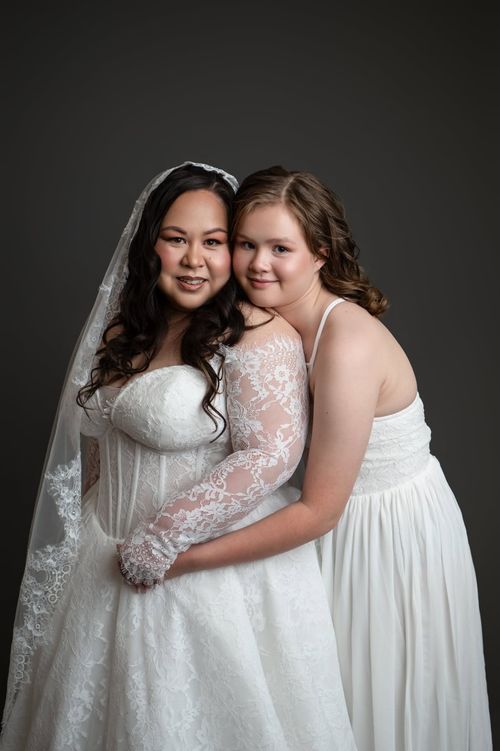 Bride in lace wedding gown and veil poses with young bridesmaid in white dress against gray backdrop.