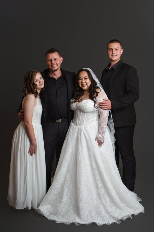 A bride in a white lace gown and veil poses with three family members in formal attire against a dark backdrop.