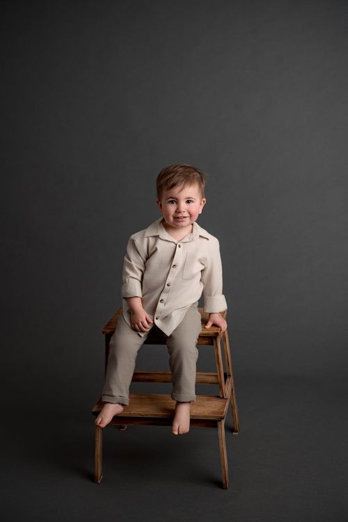 Smiling toddler boy in beige linen outfit sitting on a wooden step stool against a gray background.