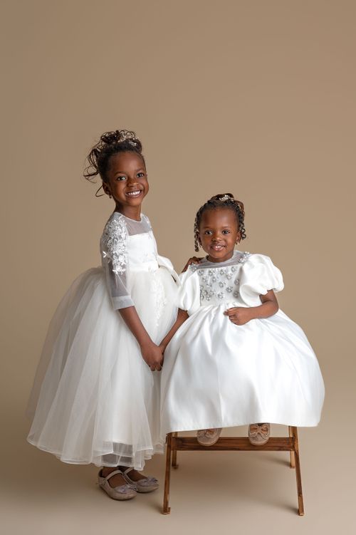 Two young girls in white flower girl dresses pose for a portrait, one standing and one seated on a wooden stool.