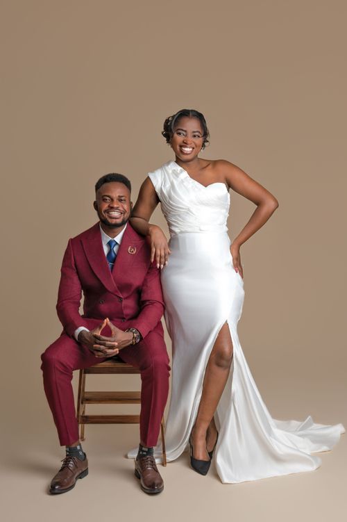 Man in burgundy suit seated beside woman in white wedding gown in elegant studio portrait.