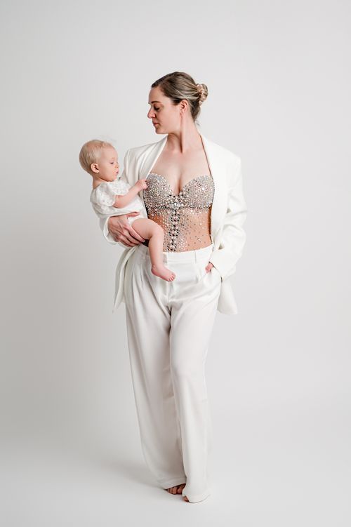 Woman in white suit and jeweled corset holds a baby against a white background.