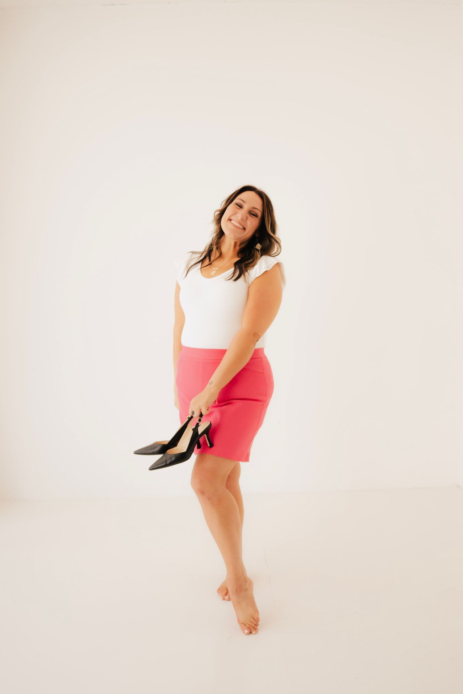 Woman in white top and pink skirt holding black heels, smiling in a bright studio setting.