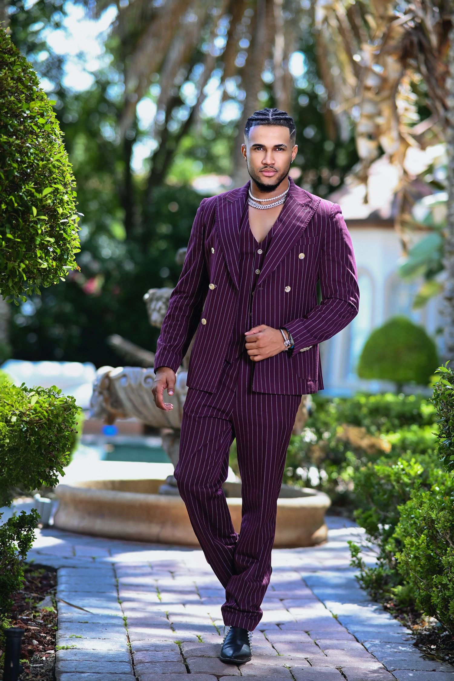 Man in purple pinstripe three-piece suit walking through lush garden with fountain and tropical greenery.