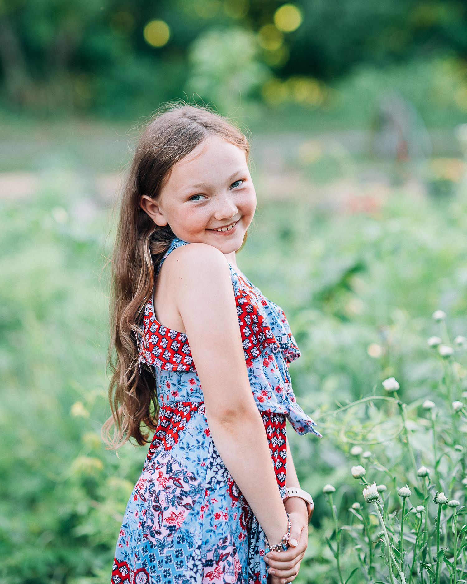 A smiling young girl in a floral dress stands among wildflowers in a lush green garden.