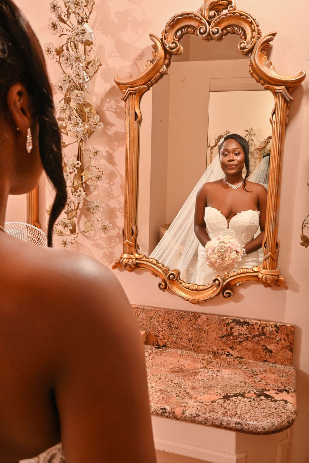 A Black bride in a strapless white gown and veil holds a bouquet, reflected in an ornate gold mirror.