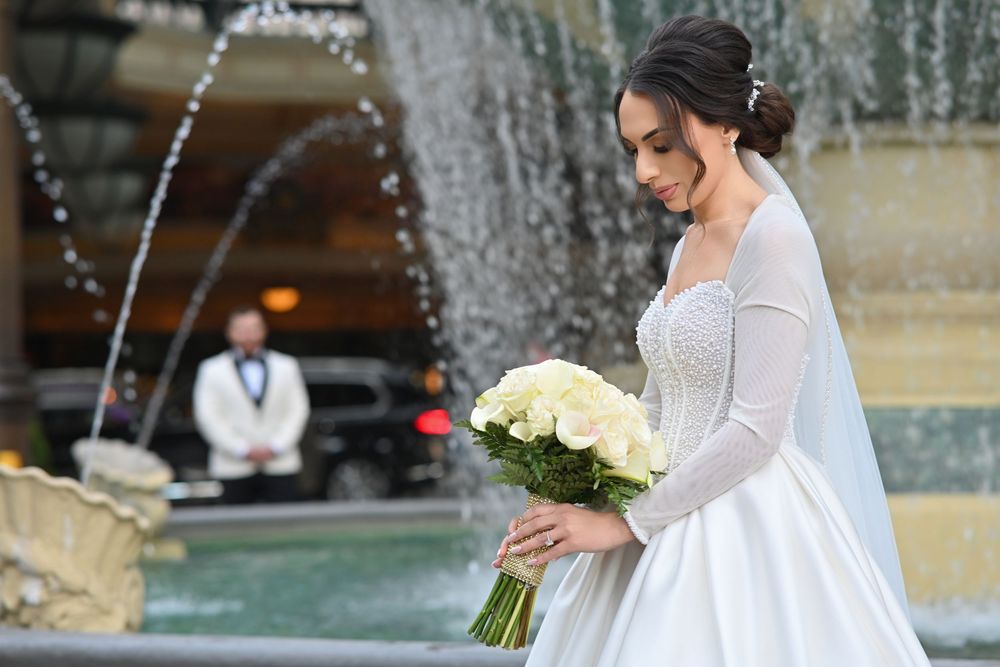 Elegant bride in white gown holding bouquet of white roses stands before ornate fountain at outdoor wedding.