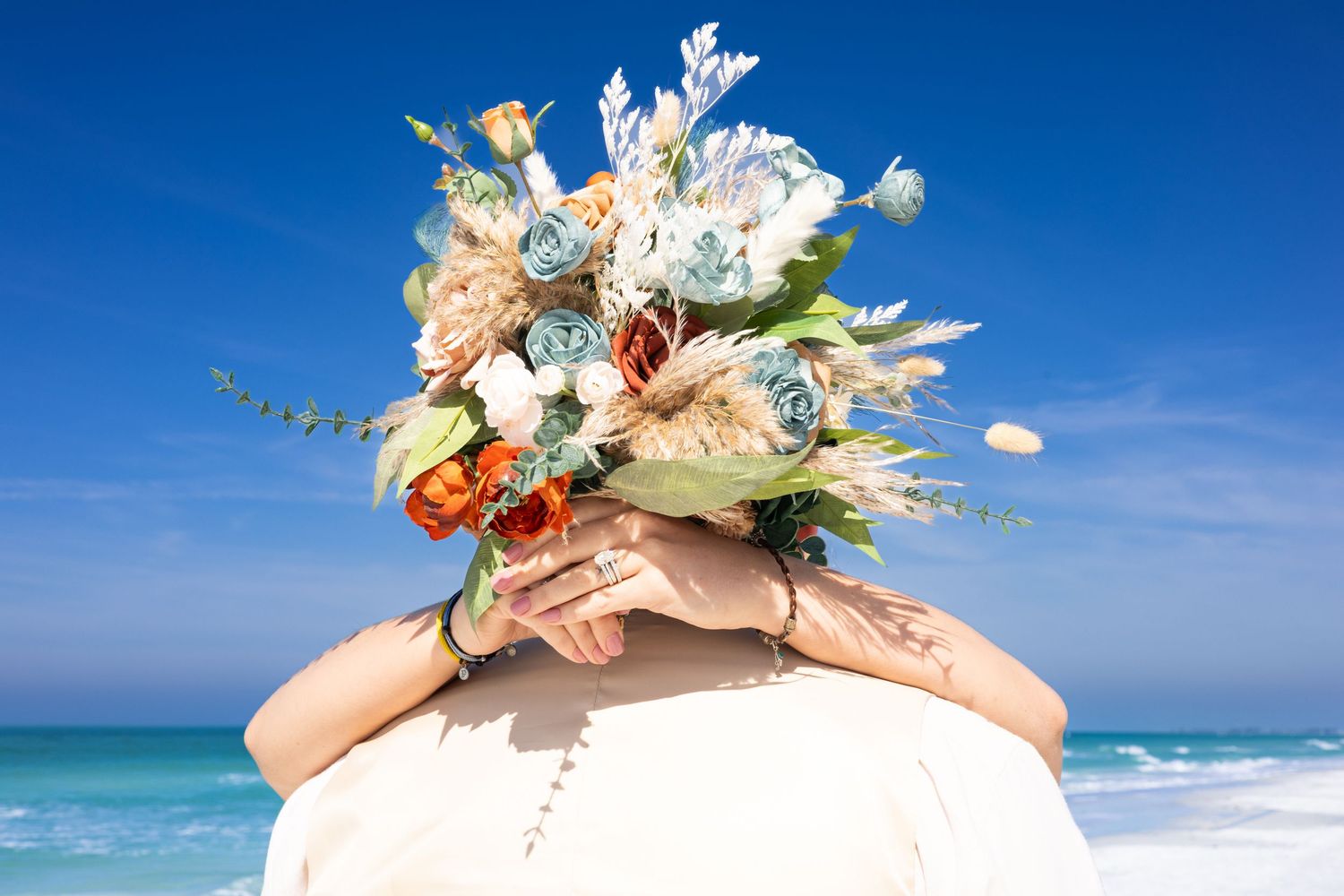 Bride holding a colorful beach wedding bouquet with blue roses and tropical flowers against a blue ocean sky.