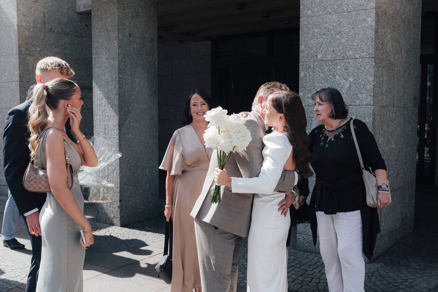 Bride in white dress receives hug from guest while bridesmaids in gray stand outside modern building on sunny wedding day.