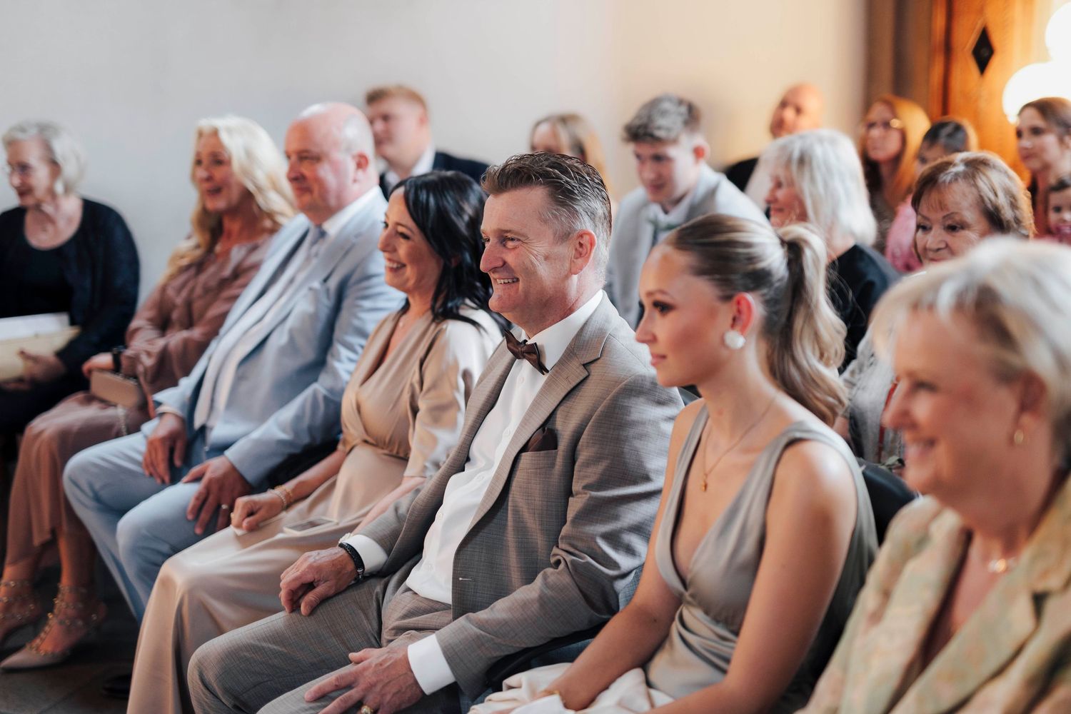 Guests smiling and laughing during a wedding ceremony, seated in rows at an elegant indoor venue.