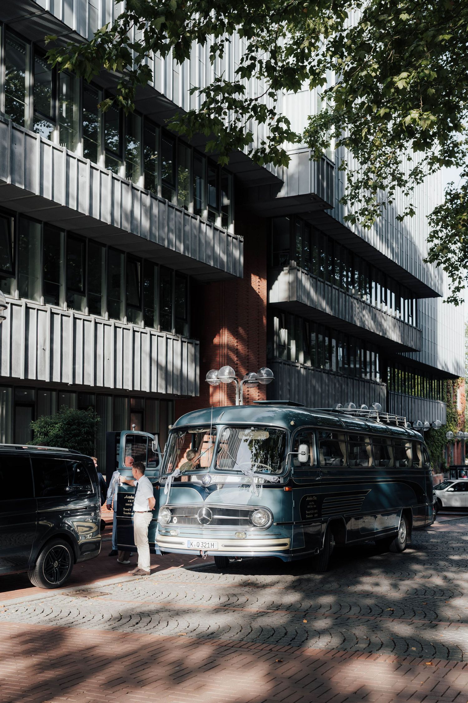 Vintage Mercedes-Benz bus parked outside a modern building, with people standing nearby on a brick sidewalk shaded by trees.