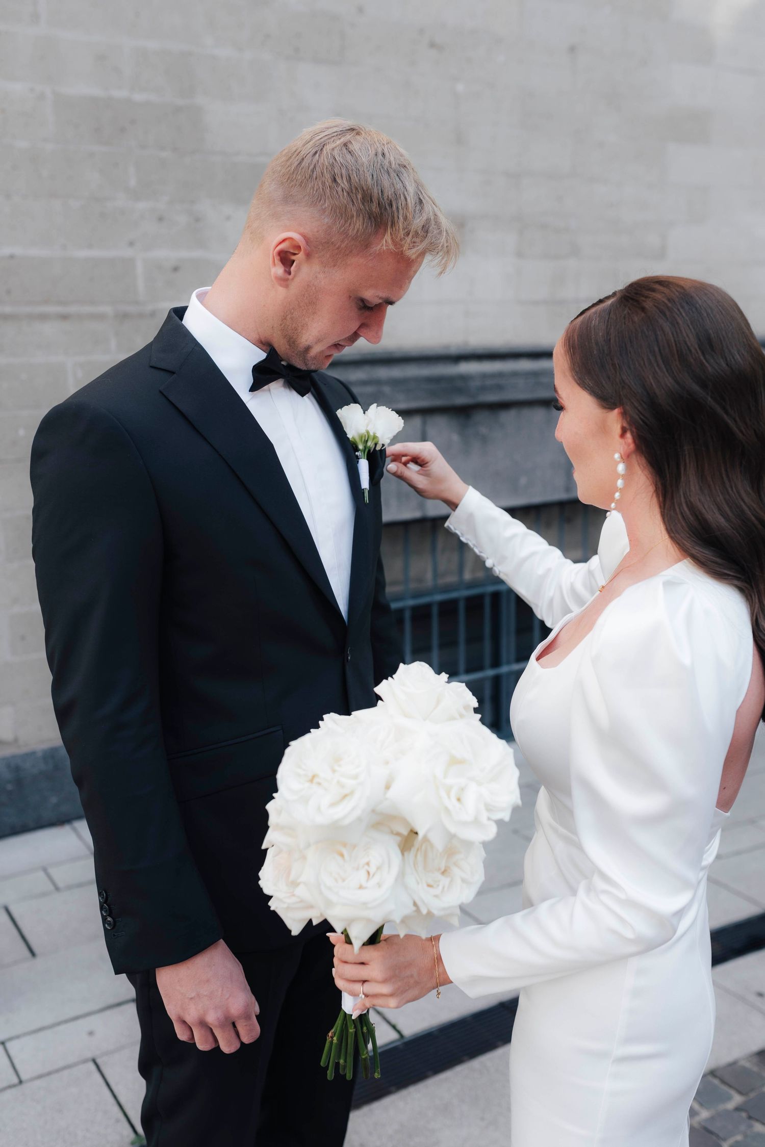 Bride in white dress adjusts groom boutonniere while holding white rose bouquet outdoors on wedding day.