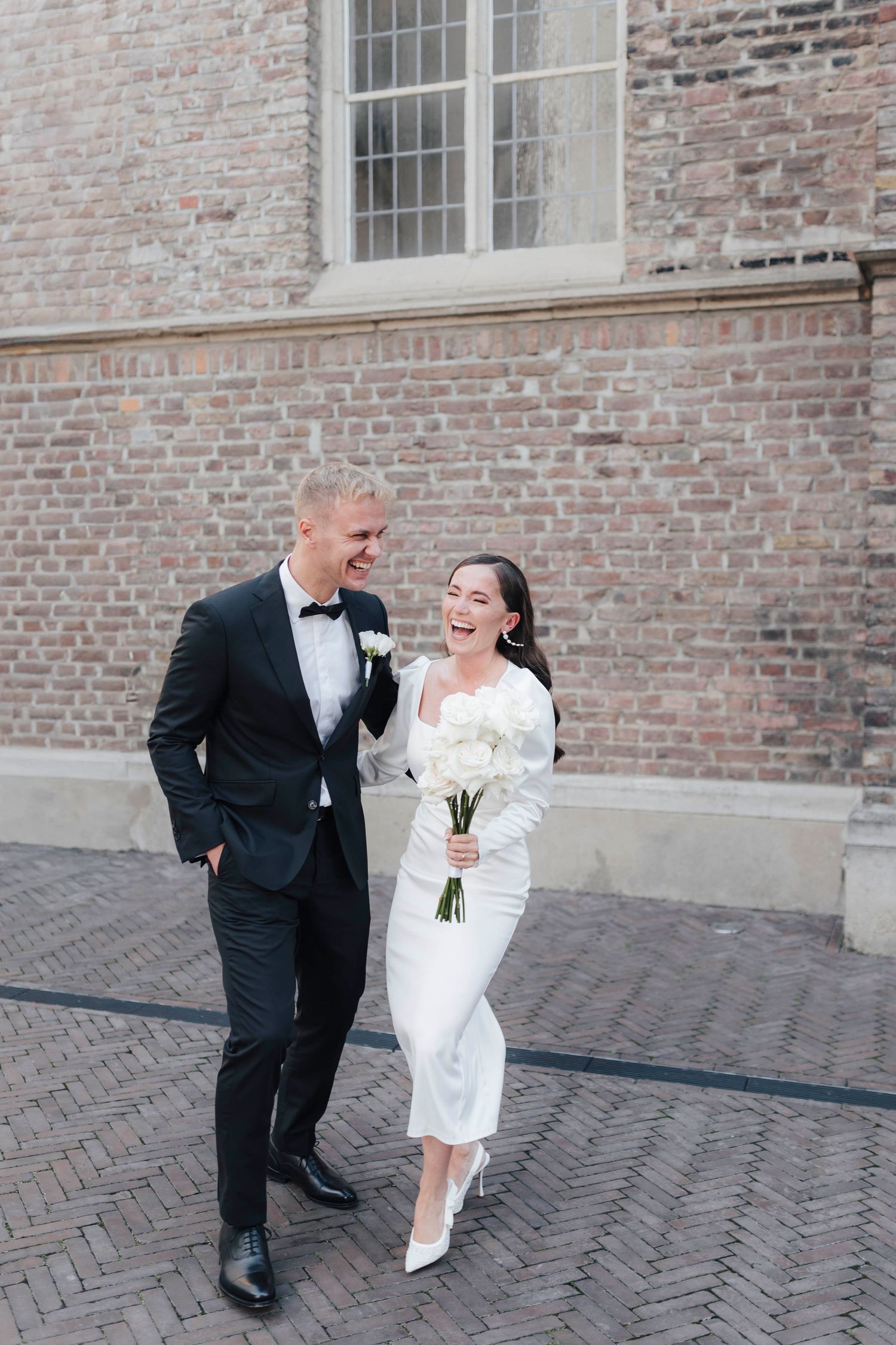 Happy bride and groom walking together on cobblestone street, bride in white midi dress holding bouquet, groom in black tuxedo.