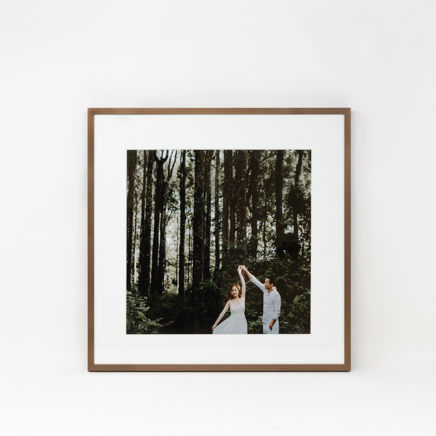 A framed square wedding photo of a couple dancing in a tall forest, displayed in a warm wood frame.