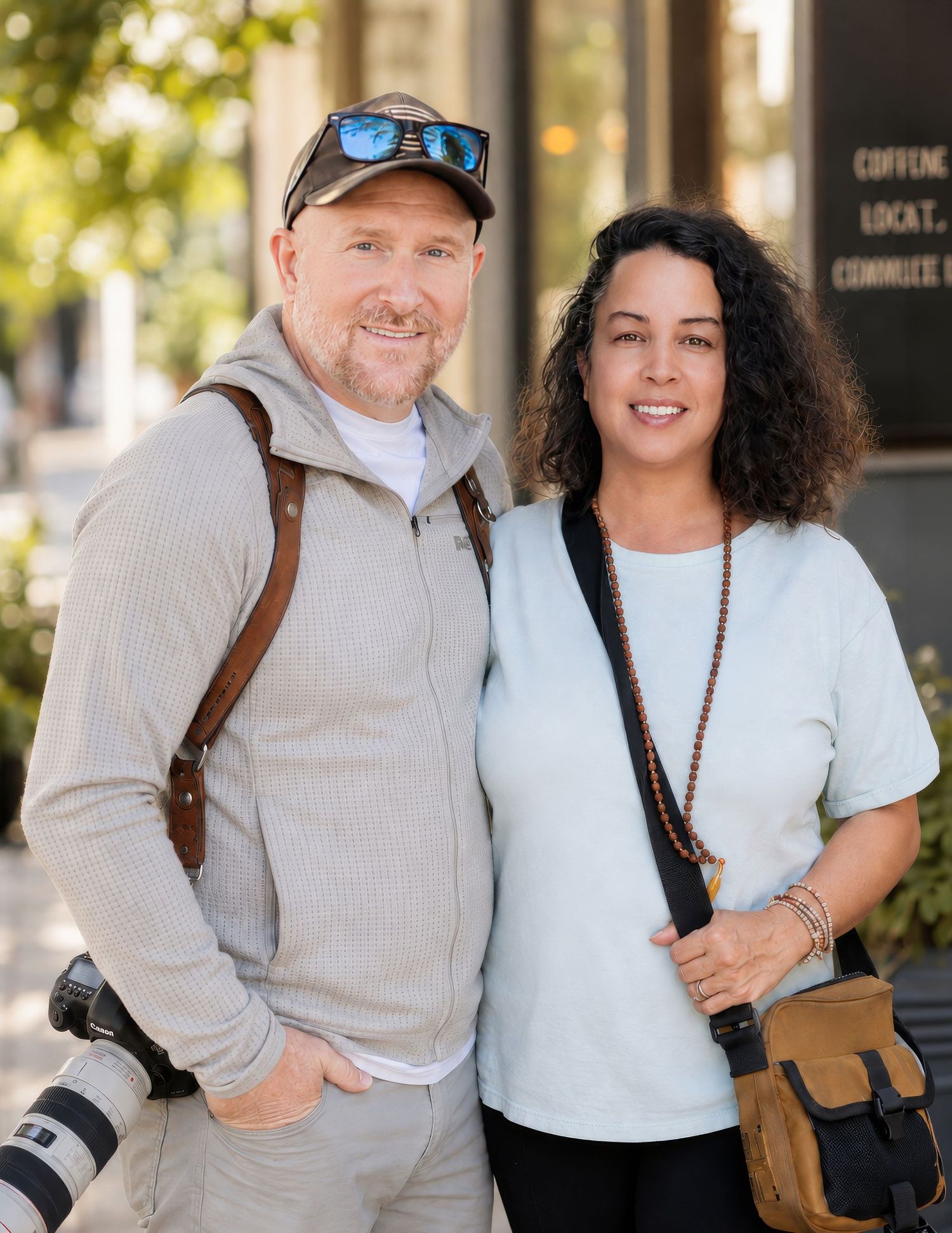 A male photographer with a camera and a woman holding a phone, both wearing crossbody bags outdoors.