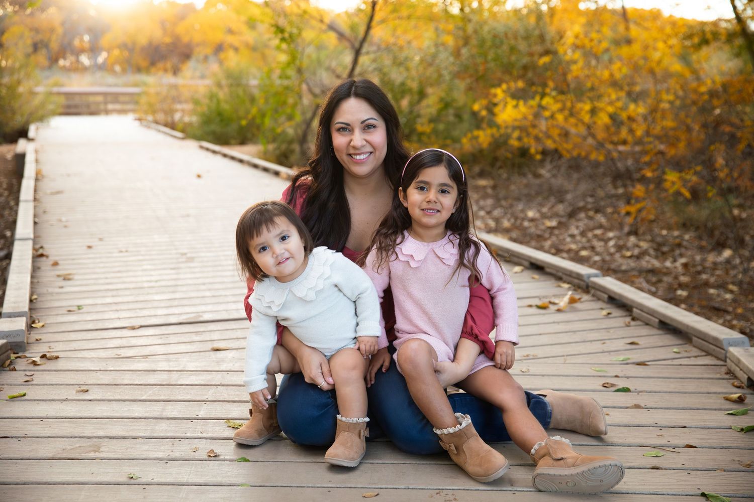 A mother sits on a wooden boardwalk with her two young children surrounded by golden autumn foliage.