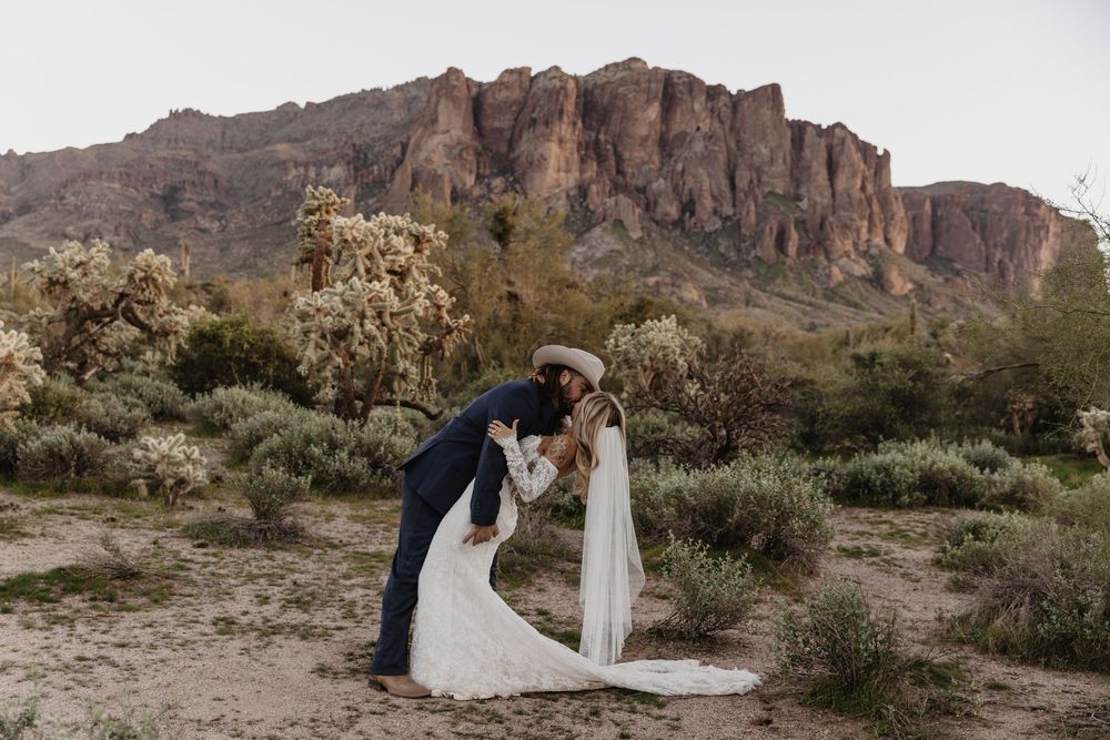 Bride and groom share a romantic kiss in the desert with rugged mountain backdrop and desert plants surrounding them.