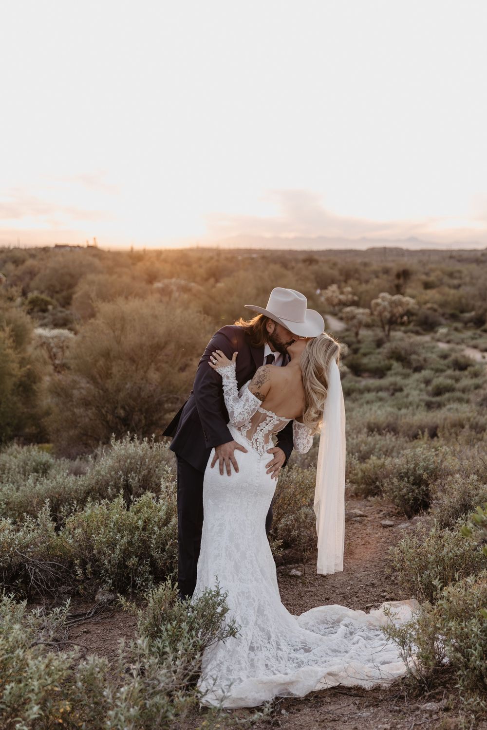 Bride and groom share a romantic kiss at sunset in a desert landscape, bride wearing lace gown and cowboy hat.