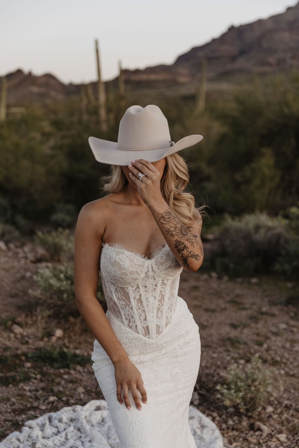 Bride in white lace corset gown and cream cowboy hat poses in Arizona desert with saguaro cacti.