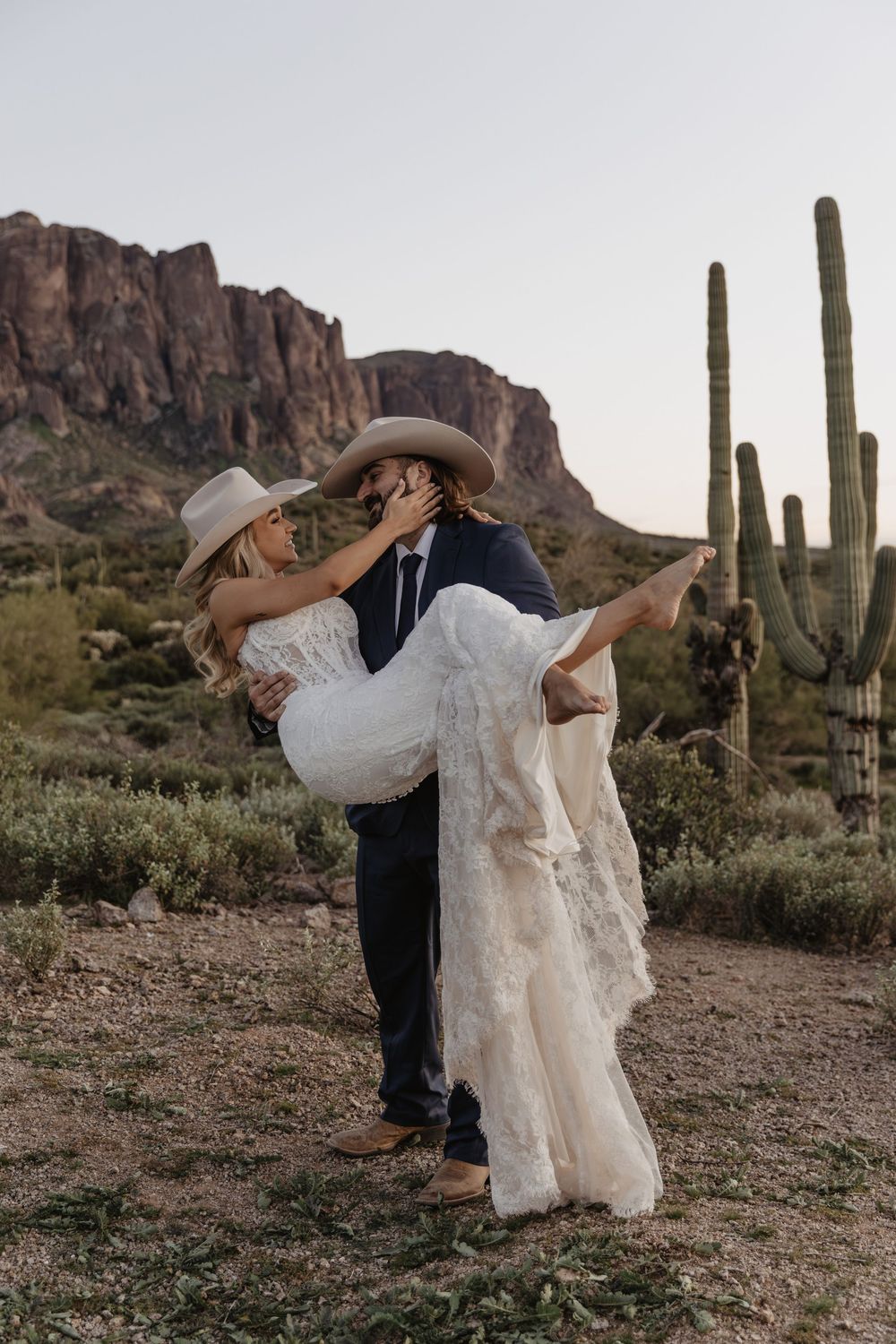 Groom lifts bride in lace dress and cowboy hats, sharing a romantic moment in Arizona desert with saguaro cacti.