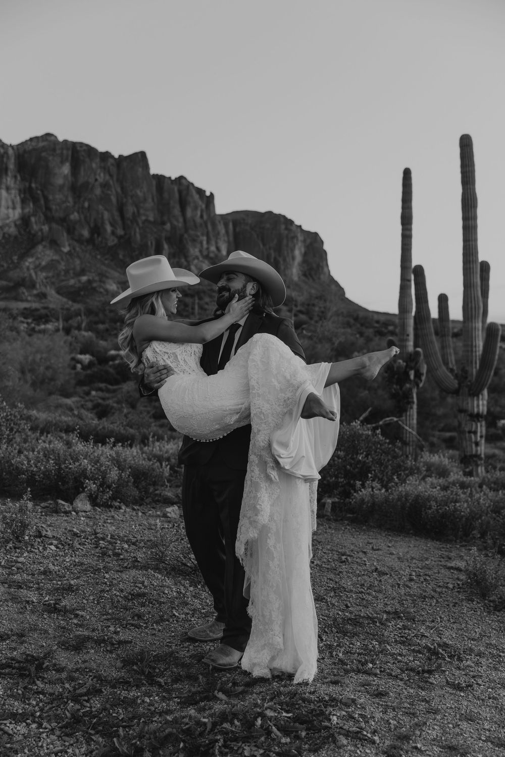 Black and white photo of a couple in cowboy hats, groom carrying bride in desert with saguaro cacti.