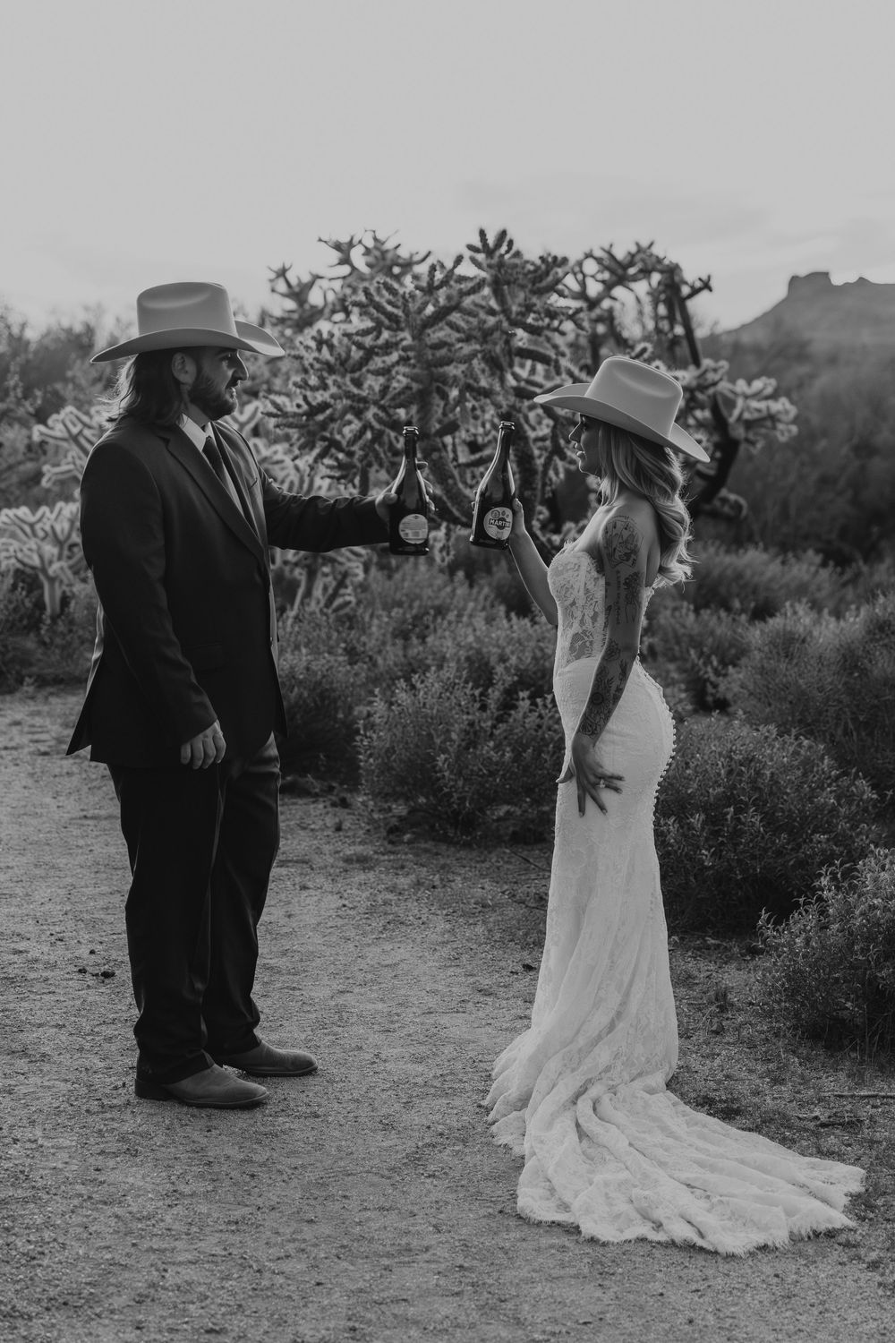 Black and white photo of a bride and groom in cowboy hats exchanging rings in a desert landscape.