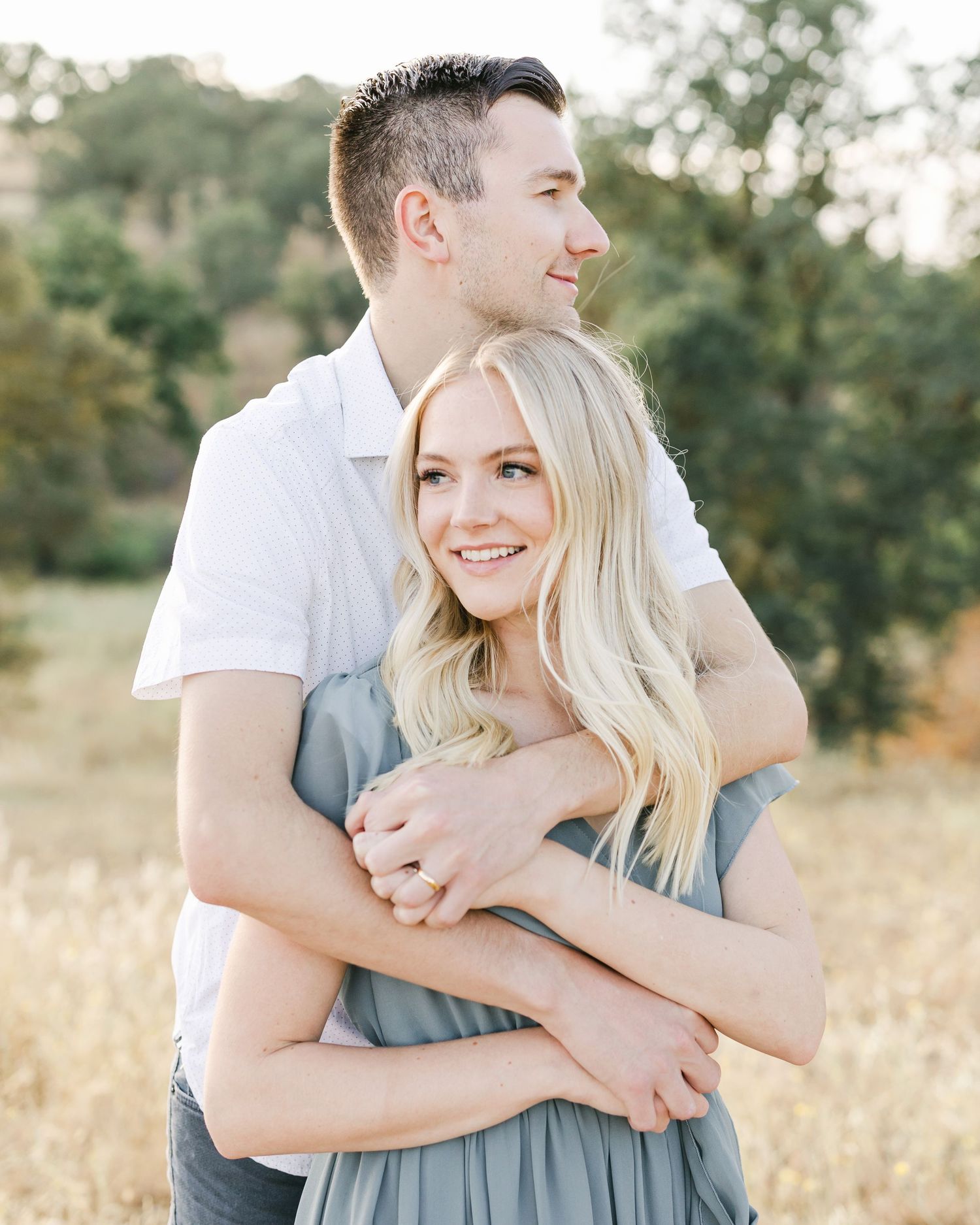 A couple embraces in a sunny field, woman in gray dress smiling, man in white shirt holding her from behind.