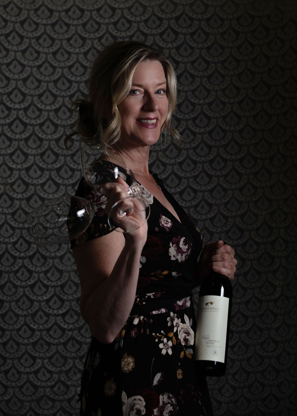 Woman in floral dress holding wine bottle and glass, smiling against decorative wallpaper background.